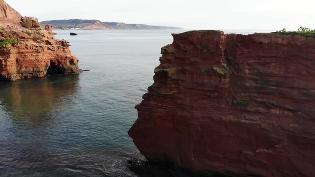 Stunning aerial drone footage of majestic red sandstone sea cliffs in Devon overlooking calm ocean waters.