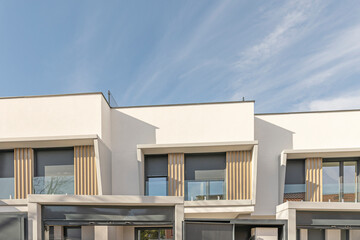 Upper facade of modern white buildings featuring stepped rooflines, balconies with vertical wooden slats, glass railings, and dark shutters under a clear sky. © Toyakisfoto.photos