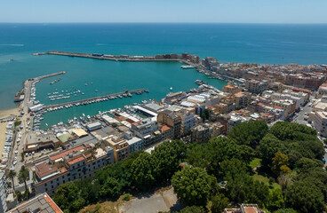 Fototapeta premium High aerial view of the harbor in Anzio, a coastal town in province of Rome, Italy. There are boats moored in turquoise sea water, coastal buildings, and the vast open horizon under a clear sky.