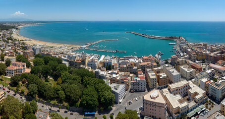 Fototapeta premium High aerial view of the harbor in Anzio, a coastal town in province of Rome, Italy. There are boats moored in turquoise sea water, coastal buildings, and the vast open horizon under a clear sky.