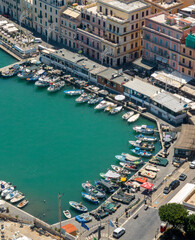 Fototapeta premium Aerial top view of a scenic coastal harbor featuring boats docked along a curved stone promenade lined with charming buildings and vibrant turquoise sea water. It's Anzio, a coastal town in Italy.