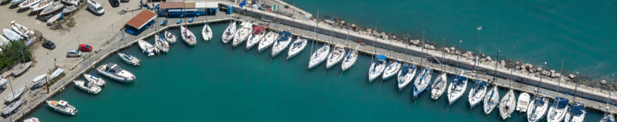 Naklejka premium Aerial panoramic view of a picturesque marina filled with numerous white boats and yachts docked along a stone pier, surrounded by clear turquoise sea water on a sunny bright day.