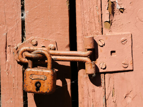 An antique latch with a heavily rusted padlock attached to a weathered wooden door painted reddish-brown.