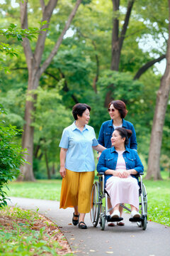 Vertical shot of Asian middle aged women walking with Asian woman with disability in wheelchair through park, supporting mobility and care. Useful for caregiving, inclusion, rehabilitation