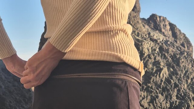 Woman zipping pocket on skirt outdoors near rocky cliff. Close-up static shot capturing the tactile motion of metal fastener closing.