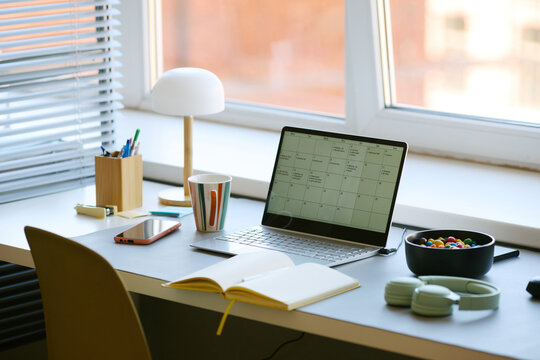Home office desk displaying laptop with calendar, notebook, smartphone, headphones, coffee mug, supporting remote work planning and daily productivity. Wide shot for hybrid work marketing