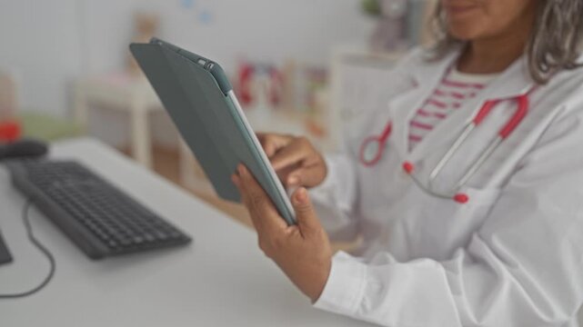 Middle age woman doctor in white coat holding tablet, touches tablet screen while seated at desk in building; care digital health.