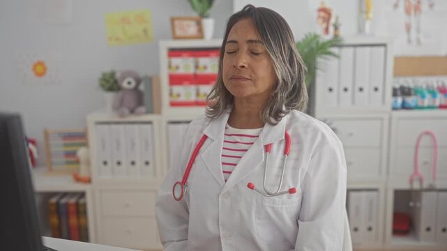 Woman doctor in white coat with pink stethoscope closing eyes and tilting head while seated at exam desk with teddy and file shelves in pediatric clinic building; fatigue recovery.