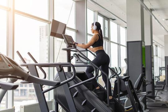 Young Black woman wearing headphones training on stair climber, improving cardiovascular endurance, leg strength and stamina during a focused workout in a bright gym
