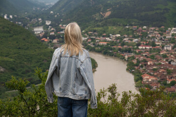 Woman standing on a hill admiring the panoramic landscape view of Mtskheta town, framed by green mountains with the Kura river winding through the valley © yaroslav1986