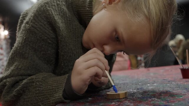 Focused blonde girl paints a small wooden slice with bright blue paint using a stick, concentrating on her art and craft project at a table