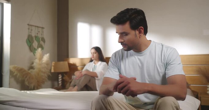 Indian young adult gen z upset male female couple pair quiet sitting on bed feel bad hurt think deep have family issue tense tired man woman look sad show distance after fight argue lost job room home