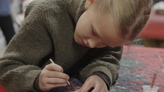 A young blonde girl focuses intently while painting a small wooden disc with red paint on a colorful, paint splattered workshop table