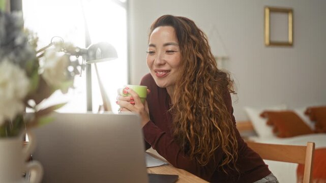 Woman holding green mug, smiling and leaning toward laptop on wooden table in studio; relaxed focus.