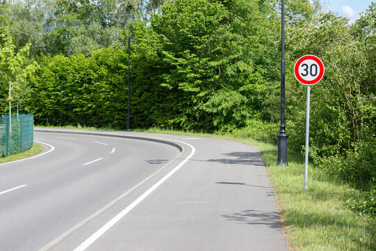 Curved road with a speed limit sign indicating 30 km/h, surrounded by lush green trees and a fence, under clear blue skies in a rural setting