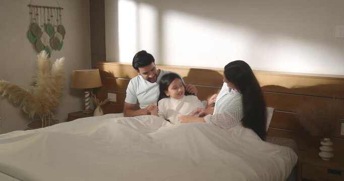 Indian man woman sitting on cozy bed do tickling to girl talk speak show love care enjoy day living room home. Young adult mom dad male female play kid child spend family time have fun joy house place