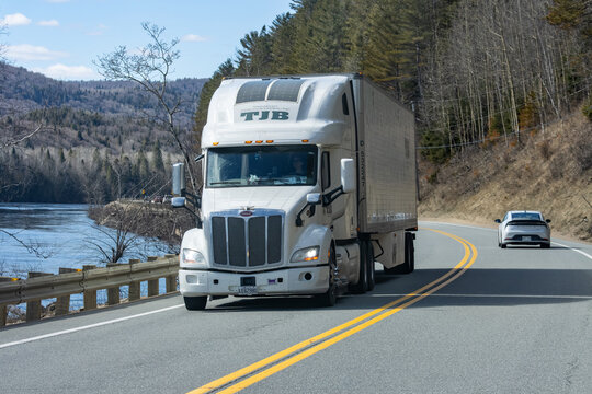 Quebec, Canada, 24 April 2026 : A white Peterbilt semi truck from TJB Transport drives on a winding road beside a river.