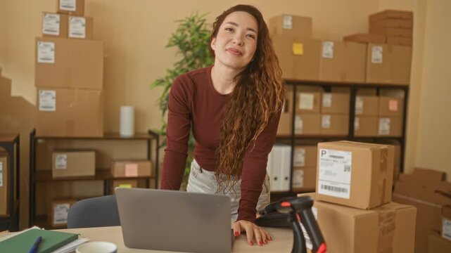 Woman leans on desk with laptop and stacked shipping parcels, hand resting on table, smiling in building office space; entrepreneurial confidence.