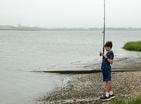 A young boy stands on a pebbled shore concentrating as he reels in a line at a calm, overcast riverbank, capturing quiet patience, outdoor recreation and a summertime hobby by the water.