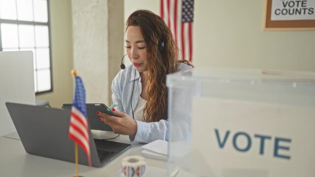 Woman holding tablet and tapping screen with hand, headset on near american flag and vote box in polling building; civic pride.