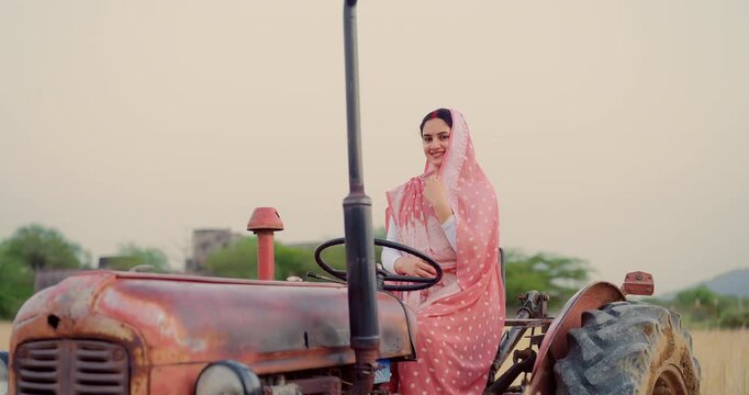 Indian young adult woman sitting on tractor learn driving steering wheel look camera at farm rural area. Happy female wear saree staring cam do casual pose enjoy desi life day village ground place