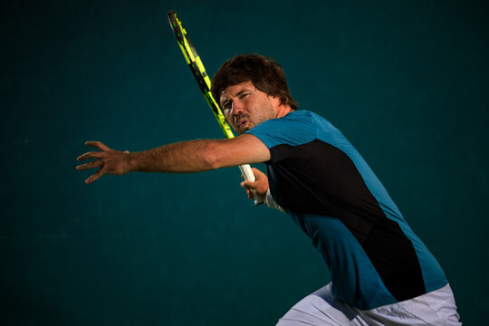 Tennis player hitting a forehand on outdoor green tennis court. Focused male athlete in action with racket and ball captured in dramatic lighting for sports advertising and editorial