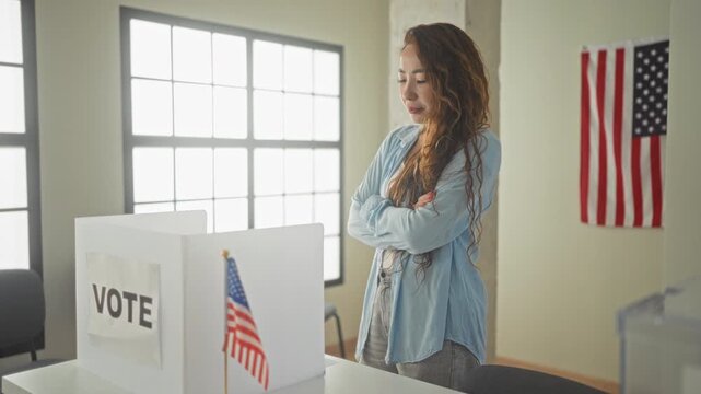 Woman at voting booth with hand to chin studying a ballot beside an american flag inside a polling building; thoughtful civic duty.