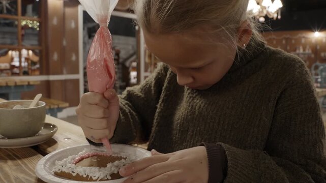 A focused young girl uses a piping bag to squeeze pink icing onto a decorated gingerbread cookie during a holiday workshop