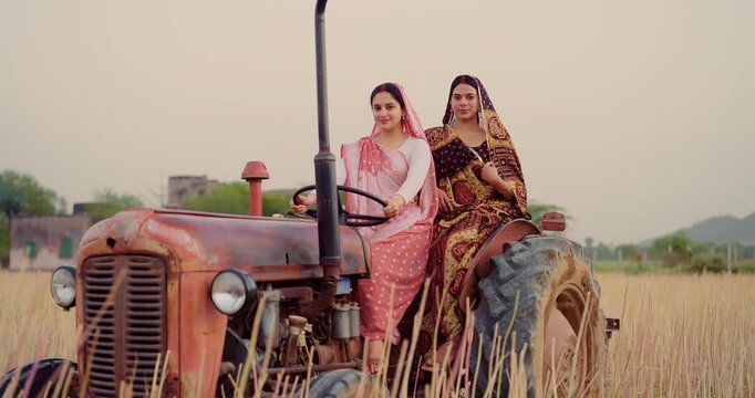 Indian young adult two woman sitting on tractor learn driving steering wheel look camera at farm rural area. Happy female wear saree staring cam do casual pose enjoy desi life day village ground place