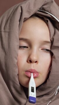 Sad little girl wrapped in a blanket measuring her temperature with a digital oral thermometer, showing symptoms of a cold, flu, or seasonal illness during a medical check up