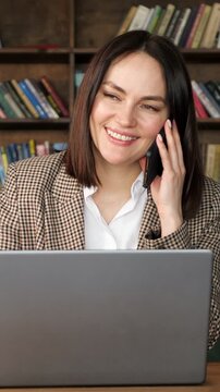 Confident business woman having a friendly conversation on her smartphone while working on a laptop computer, sitting at a desk in a modern library or a classic office interior