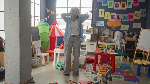 Woman teacher holding head with both hands beside a red activity table and alphabet wall amid scattered toys and play tent in a preschool classroom building; overwhelm resilience.