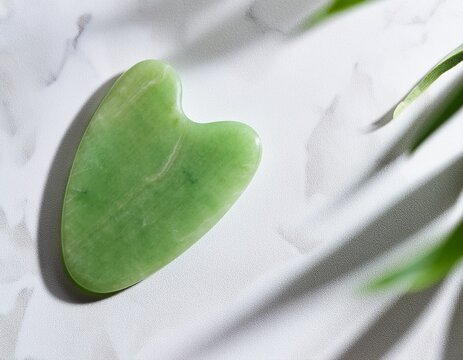 green jade gua sha massager positioned on white marble background showcasing natural textures with soft green hues and ample copy space for text