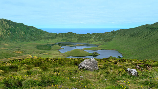 Green caldera mountain surrounds calm volcanic crater lake on scenic Corvo island, Azores, Portugal. Natural geological lagoon displays serene water landscape under cloudy sky weather.