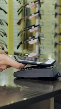 Close up of a female hand using a smartphone for a contactless payment at an optical store, with rows of eyeglasses displayed on racks in the blurred background of the shop