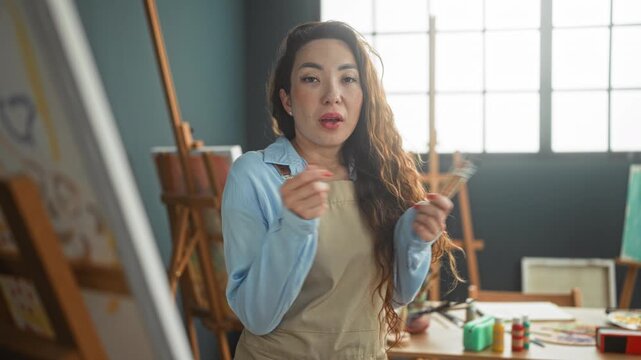 Woman painter wearing an apron holds a handful of paintbrushes and inspects a small brush by the canvas near a large window in an art studio; focused creativity.