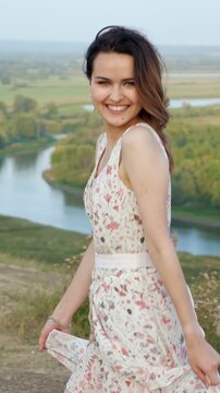 Beautiful young woman wearing a long floral dress dancing on top of a hill overlooking a river valley, her hair blowing in the wind as she spins and smiles with joy and freedom