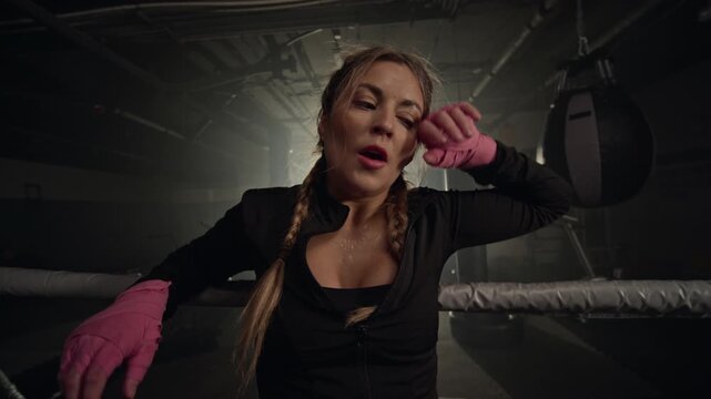 Young Caucasian woman leaning on boxing ring ropes, unwrapping boxing hand wraps and wiping sweat after workout. Ideal for fitness branding, sports motivation videos, and health campaigns