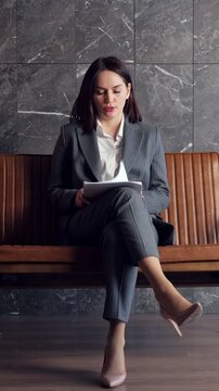Young businesswoman in a formal suit sitting on a bench, anxiously waiting for a job interview or meeting, nervously fanning herself with documents due to stress and heat in the office hall