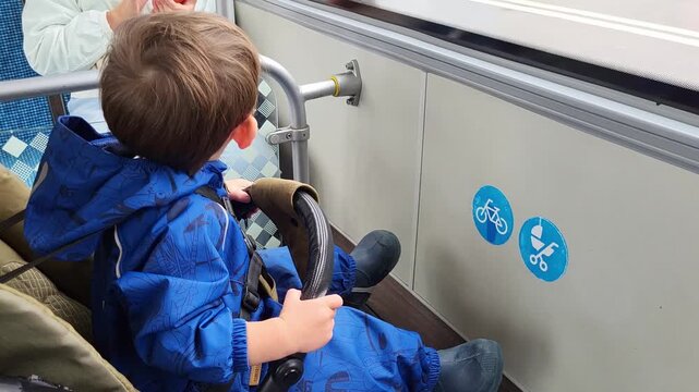 Child sitting in stroller on public transport. Little boy looking through window while riding bus. Daily commute and cityscape exploration. Transportation and urban travel concept.