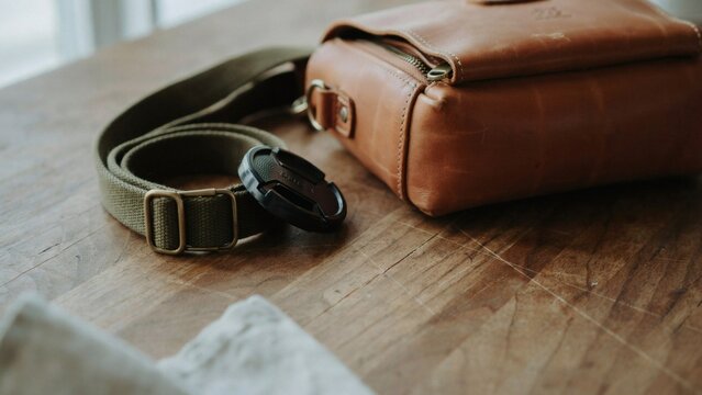 Vintage leather camera bag resting beside green fabric strap and lens cap on wooden table with weathered wooden surface strap circular black