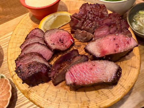 Overhead close-up of assorted grilled beef tongue cuts with glossy pink interiors, pepper seasoning and a lemon wedge.