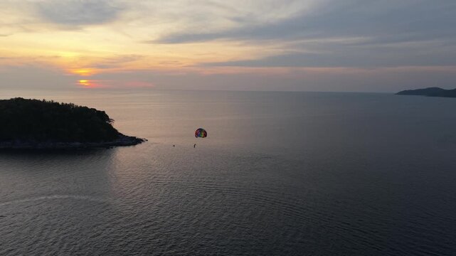 A colorful parachute floats above the ocean as a person enjoys parasailing during sunset, with a serene tropical island and calm waters creating a picturesque backdrop