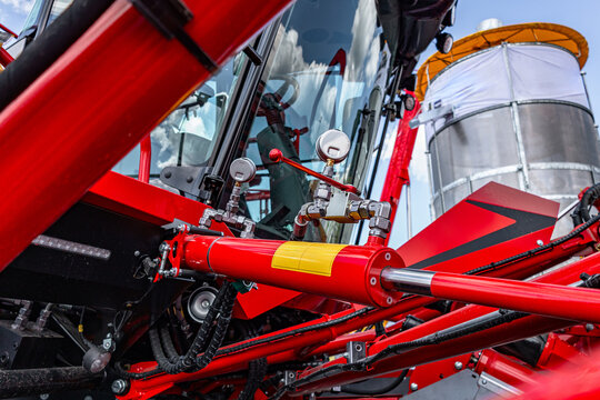 Agricultural machinery hydraulic cylinder mechanism with gauge. Modern farm equipment crop harvesting section on red metal frame with warning sticker and engineering technical details view.