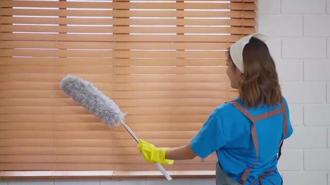 Woman cleaning wooden blind near window using gray duster with yellow glove, home housework dust cleaner in bright indoor room white brick wall tidy calm scene
