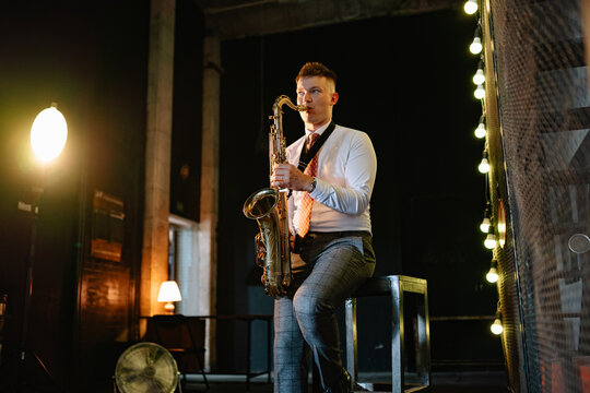 Man plays saxophone live music performance on a stage at night inside a cozy bar under warm lighting, dressed in a crisp white shirt