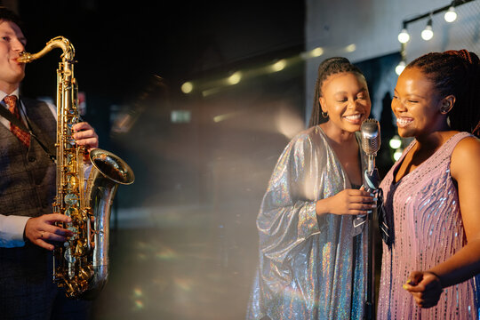 Male saxophonist performs with two joyful Black women vocalists wearing shimmering dresses, singing into a vintage microphone at a dimly lit industrial bar during an evening concert