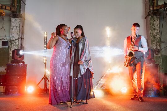 Two Black women sing and a man plays saxophone on a brightly lit stage, creating a vibrant, golden-hued live jazz performance for an audience