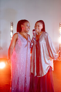 Two talented women stand side by side on a stage and sing a song into a shared vintage microphone while wearing shiny dresses during an indoor evening performance illuminated by warm stage lights