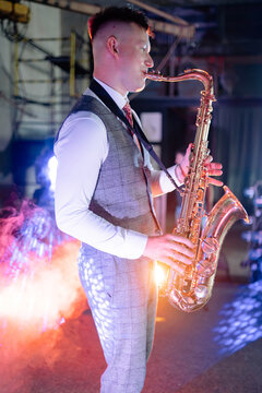 Professional male musician playing a shiny brass saxophone during a live jazz performance on an industrial stage lit by bright spotlights and filled with atmospheric stage smoke at night